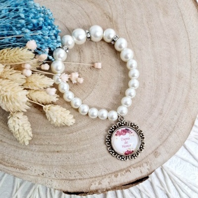 Pulseira de pérolas brancas com pendente redondo metálico com texto floral sobre base de madeira clara e flores secas.