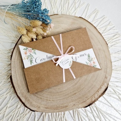 Envelope de convite em papel kraft com decoração floral e fita rosa sobre base de madeira