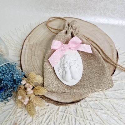 Saco de tecido castanho claro com laço rosa e medalha de anjo branco sobre madeira e flores secas ao lado.