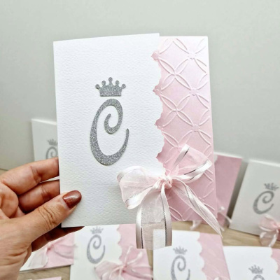 Convite branco e rosa com laço branco e inicial C em glitter prateado, fundo com outros convites similares