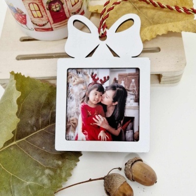 Enfeite branco em forma de moldura com laço e foto de mulher e criança, bolotas e folha seca ao lado