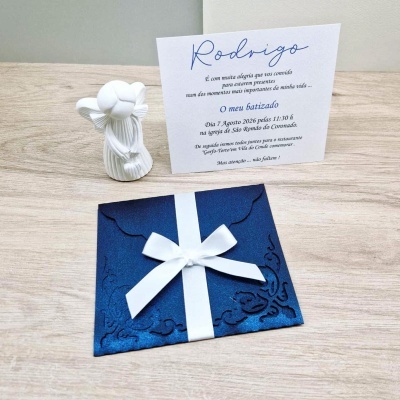 Convite batizado papel azul com laço branco e cartão com texto ao lado, ao lado uma estatueta de anjo branca sobre mesa de madeira.