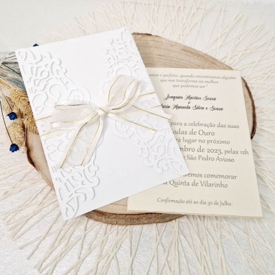 Convite de casamento branco com laço dourado e cartão com texto em papel creme sobre base de madeira