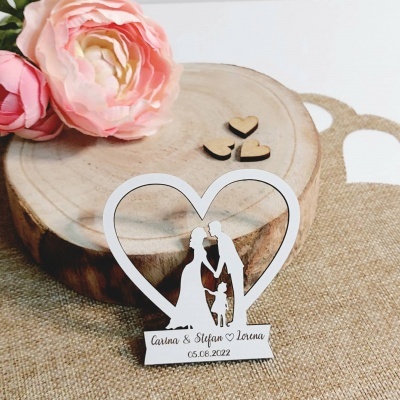 Decoração branca de casal e criança em forma de coração com data, sobre tronco de madeira e flores cor-de-rosa.