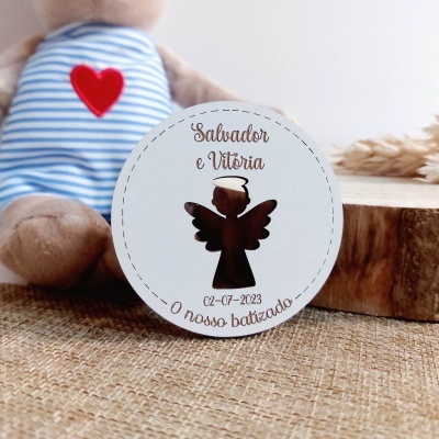 Etiqueta redonda branca com anjo recortado e texto para batizado, junto a peluche com camisola azul às riscas e coração vermelho