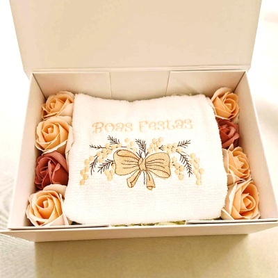 Toalha branca bordada com texto Boas Festas em caixa com flores artificiais.