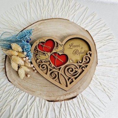 Anel de porta em madeira em forma de coração com nomes gravados e anéis, sobre base de madeira e flores secas.