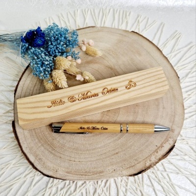 Estojo e caneta de madeira clara gravados com nomes, sobre madeira redonda decorativa com flores secas.