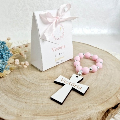 Caixa branca com fita rosa e terço rosa com cruz branca nome Victória sobre tronco de madeira
