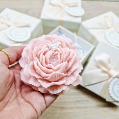 Vela decorativa rosa em forma de flor com caixas de presente brancas ao fundo.