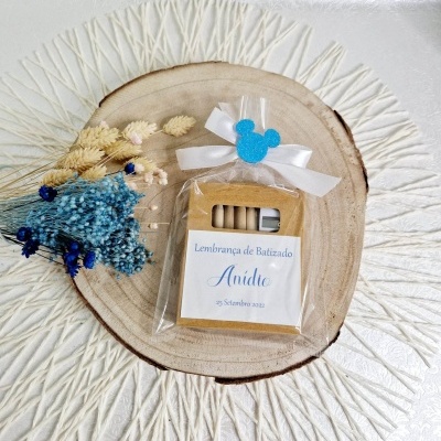 Lembrança de Batizado com lápis em embalagem plástica com laço branco e detalhe azul, sobre base de madeira e tapete branco texturizado com flores secas.