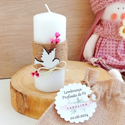 Vela branca decorativa com pássaro de madeira e flores secas, colocado sobre suporte de madeira, com etiqueta e tecido ao lado.
