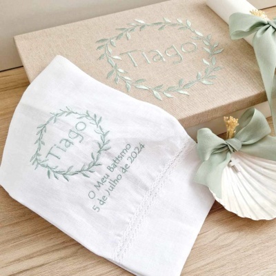 Toalha branca bordada com nome Tiago e data de batismo, caixa de tecido bege com bordado similar, concha decorativa com laço verde