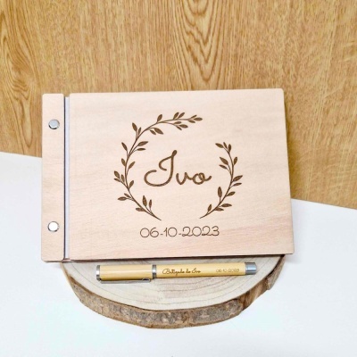 Livro de madeira clara personalizado com nome e data e uma caneta dourada num fundo de madeira e superfície branca.