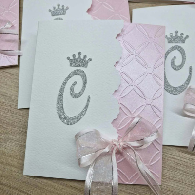 Cartões de convite brancos e rosa com letra C prateada, coroa e laço de fita em madeira
