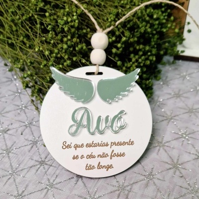 Adorno redondo branco com texto e asas verdes pendurado num cordão com duas contas sobre tecido prateado com padrão geométrico