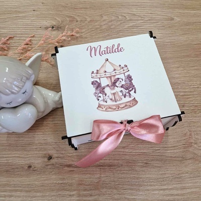 Caixa de presente branca com nome Matilde e carrossel rosa, ao lado de figura de unicórnio cerâmico sobre mesa de madeira