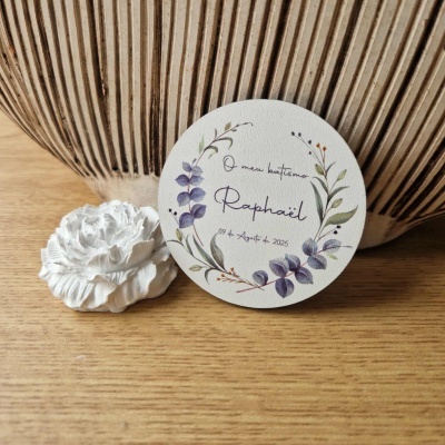 Etiqueta redonda branca com desenho de ramos e texto sobre baptismo, ao lado de flor branca em vaso de fibra