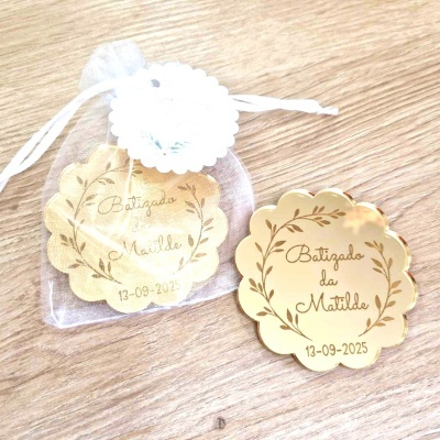 Lembrancinhas douradas redondas para batizado com texto, uma dentro de saco branco translúcido sobre mesa de madeira.