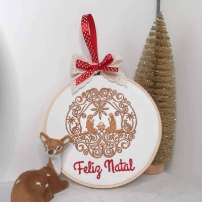 Quadro de tecido bordado com presépio dourado e texto 'Feliz Natal' em vermelho, com decoração natalícia ao lado