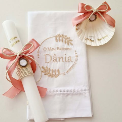 Conjunto de batismo com vela branca, toalha branca bordada e concha decorativa com laços cor de rosa.