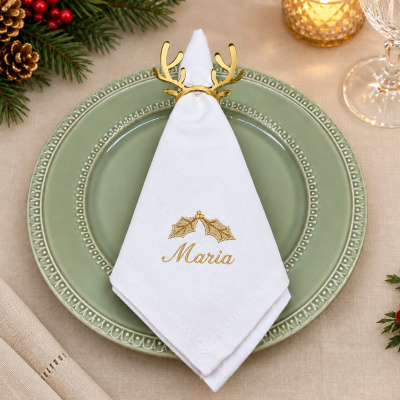 Guardanapo branco bordado com nome Maria e argola dourada em prato verde decorativo numa mesa festiva.