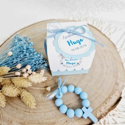 Caixa decorativa branca e azul com etiqueta redonda e terço azul com cruz sobre madeira e flores secas