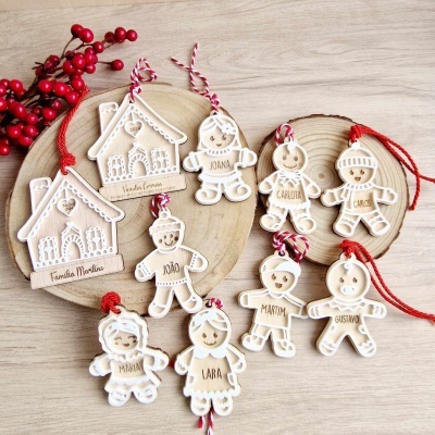 Enfeites de Natal de madeira clara com detalhes brancos e nomes gravados, pendurados em fios vermelhos e brancos, sobre bases de madeira clara com bagas vermelhas.