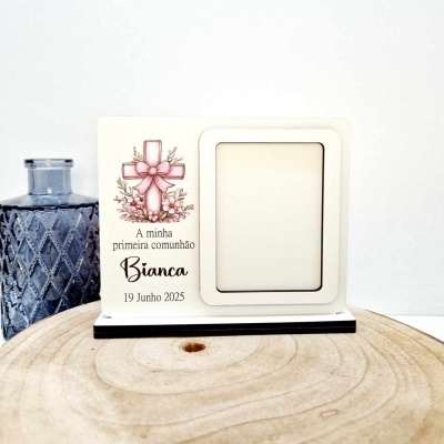 Porta-foto branco para primeira comunhão com cruz e texto, sobre superfície de madeira com jarra azul ao lado.