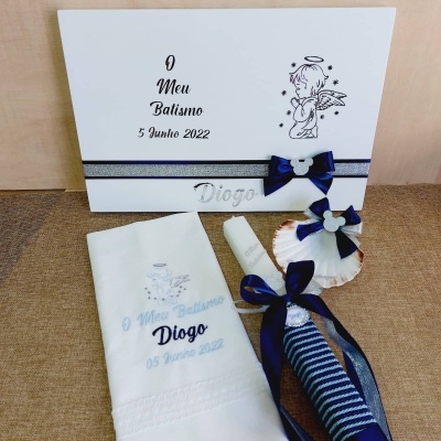 Conjunto de batismo com toalha, vela e concha decorada, e caixa branca com texto e desenho de anjo.