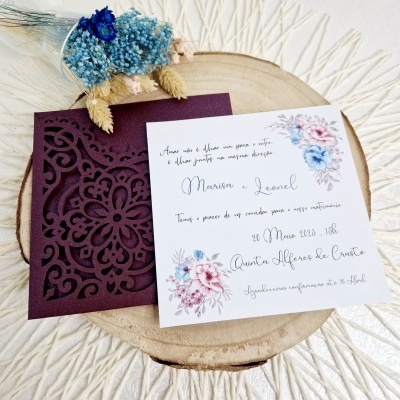 Convite de casamento em papel branco com envelope roxo e flores, sobre base de madeira com flores secas