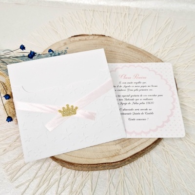Convite branco com fita rosa e coroa dourada sobre base de madeira com cartão impresso