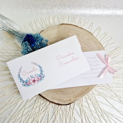 Convite de primeira comunhão branco com flores e texto cor-de-rosa sobre base de madeira
