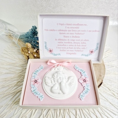 Caixa branca aberta com medalhão branco de anjo em tecido rosa e decoração floral