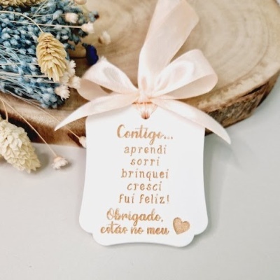 Etiqueta branca com texto dourado pendurada numa fita rosa clara com laço, colocada sobre madeira e flores secas.