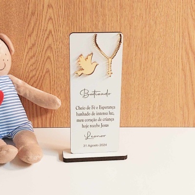 Decoração de batizado com colar dourado e pendente em forma de pomba sobre suporte branco com texto, junto a boneca de tecido vestindo camisa azul com riscas brancas