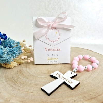 Caixa branca com laço rosa, terço cor-de-rosa e crucifixo com nome Victória sobre madeira