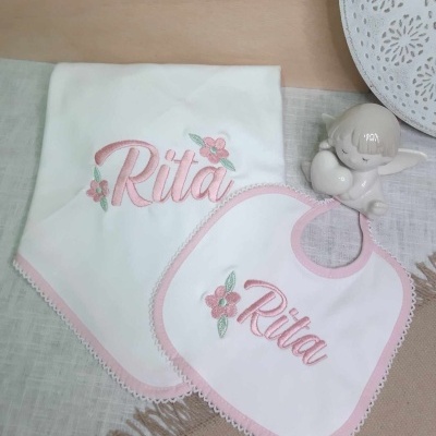 Fralda e babete bordados com o nome Rita em rosa e flores, junto a anjo decorativo e prato branco