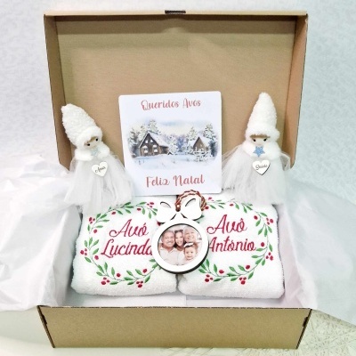 Caixa de presente com toalhas bordadas, moldura de foto, cartão natalício e figuras decorativas brancas.