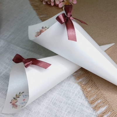 Dois cones de papel branco com fita castanha em laço e etiquetas florais sobre tecido claro
