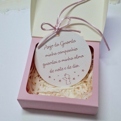 Ornamento redondo branco com cordão rosa e texto, dentro de caixa rosa com enchimento branco.