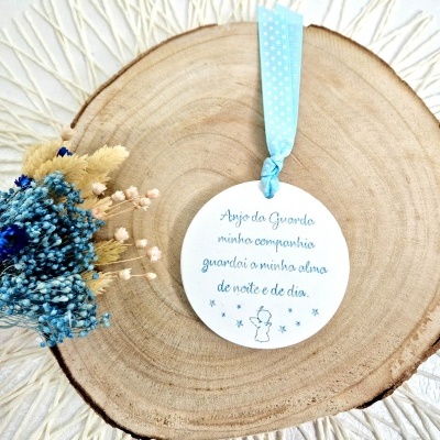 Ornamento redondo branco com fita azul e texto em português sobre base de madeira e flores secas.