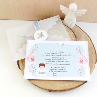 Convite azul claro com flores rosa e verde, envelope transparente com fecho de pássaro dourado, base de madeira redonda e figura decorativa branca de anjo