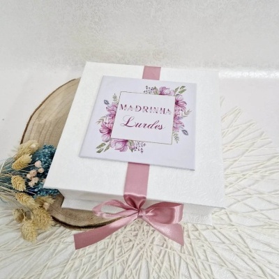 Caixa branca com fita cor-de-rosa e etiqueta floral 'MADRINHA Lurdes' sobre superfície decorativa branca e madeira, com flores secas ao lado