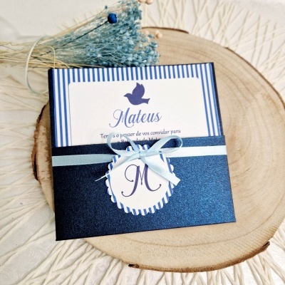 Convite de convite azul escuro e branco com nome Mateus em riscas azuis, etiqueta com M e laço azul claro num fundo de madeira clara e flores secas azuis