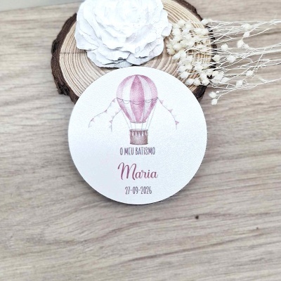 Etiqueta redonda branca com balão rosa e texto de batismo sobre base de madeira