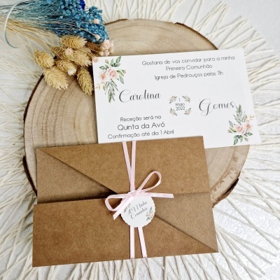 Convite para Primeira Comunhão com envelope castanho e fita rosa sobre superfície de madeira e tecido branco
