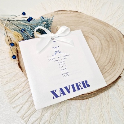 Saco presente branco com nome XAVIER e cruz de texto, sobre base de madeira e flores secas azuis.