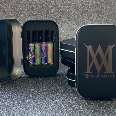 Estojo metálico preto com quatro frascos pequenos de perfume coloridos e tampa com letras douradas Manuel Altardi