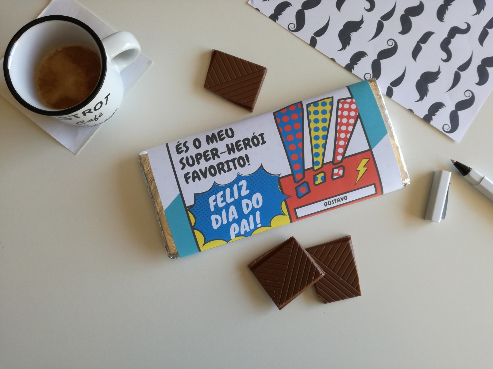 Barra de chocolate com embalagem colorida e texto de Dia do Pai ao lado de chávena de café e pedaços de chocolate